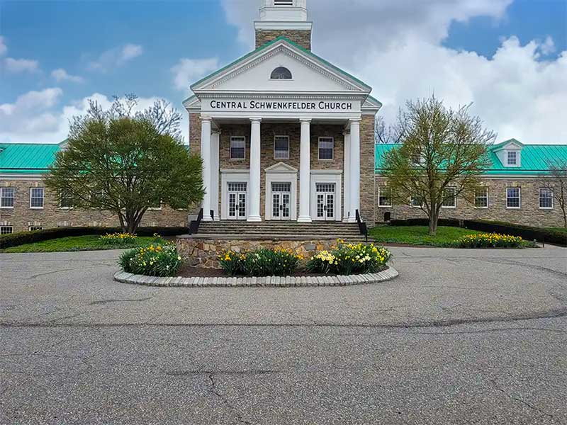The Central Schwenkfelder Church, a large brick building with white columns and green roofs, is surrounded by trees and yellow flowers, under a partly cloudy sky.