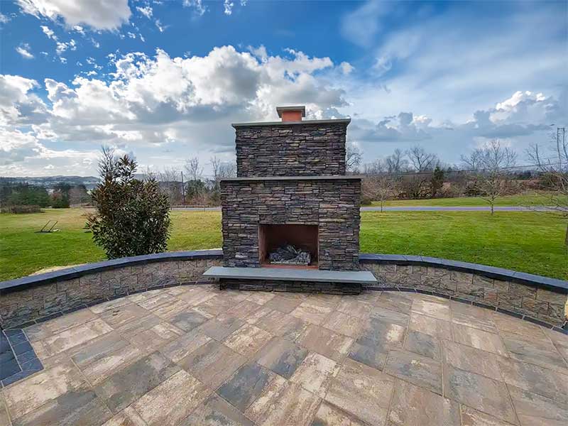 An outdoor stone fireplace sits on a tiled patio, with a grassy lawn, trees, and a cloudy blue sky in the background.