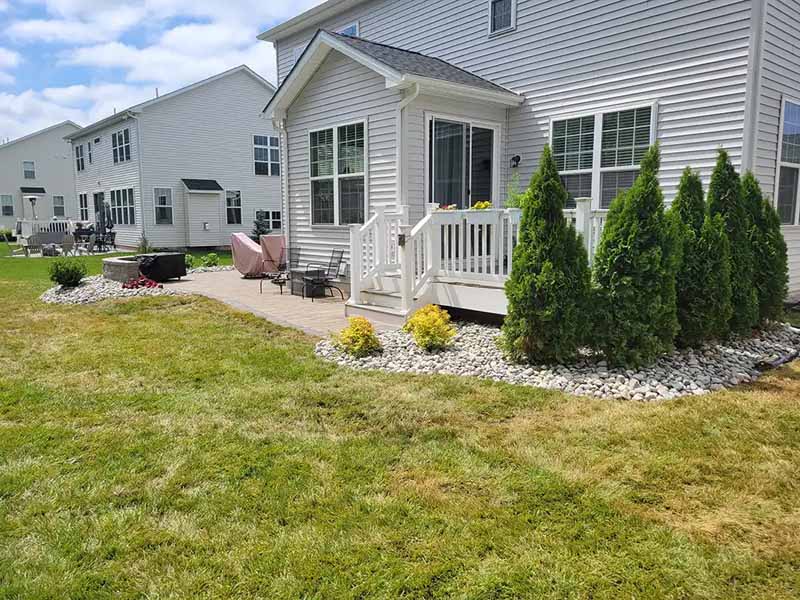 A backyard with a small porch, patio furniture, white railing, neatly trimmed bushes, and landscaping rocks next to a white house on a sunny day.