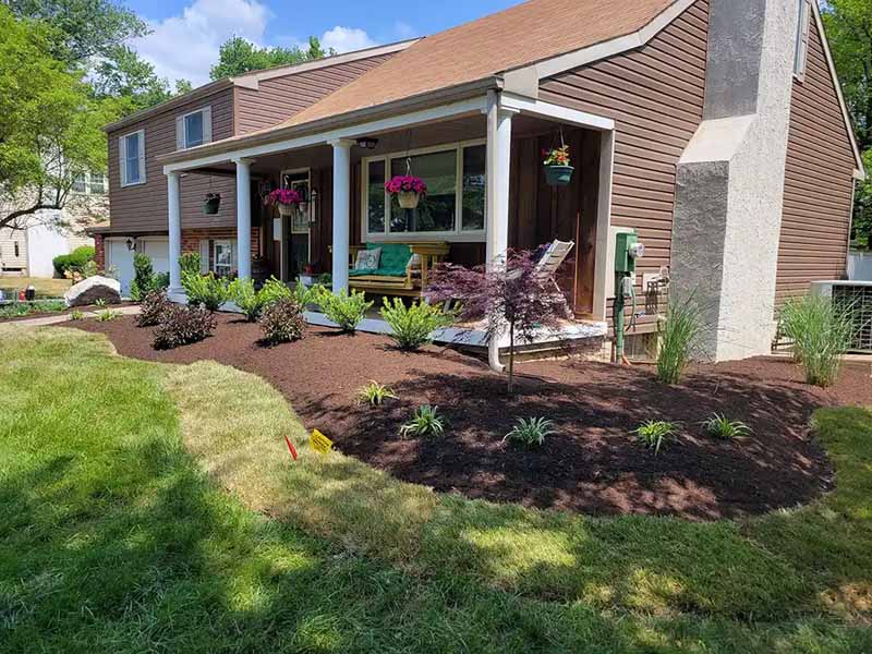 A landscaped front yard with fresh mulch, small green plants, a red tree, hanging flower baskets, a porch swing, and a brown house with white trim under a blue sky.