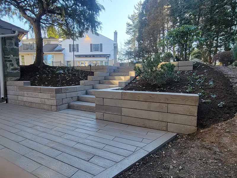 A landscaped backyard with stone retaining walls, stairs, and a patio. Freshly planted garden beds surround the steps, leading up to a white two-story house in the background.