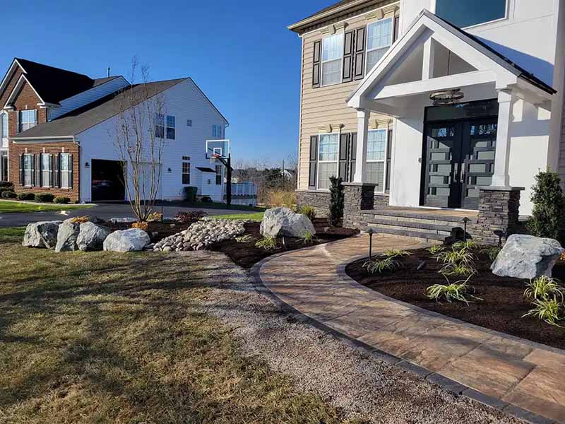 A curved stone walkway leads to a modern house with tall windows, neatly landscaped with rocks, plants, and mulch. Another house and a basketball hoop are visible in the background under a clear blue sky.