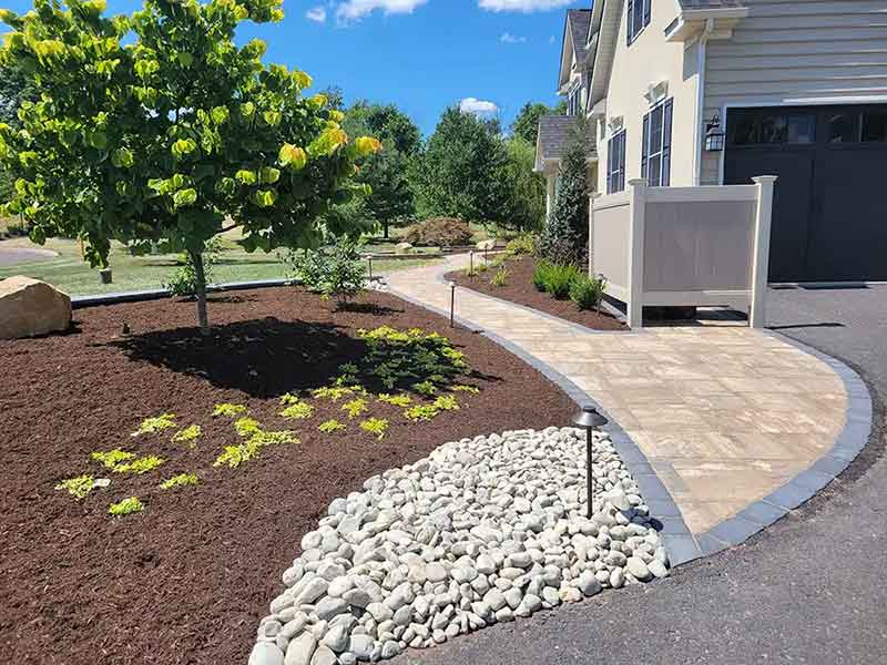 Curved stone path beside a house with mulch, small green plants, a tree, and a section of white decorative rocks under a bright blue sky.