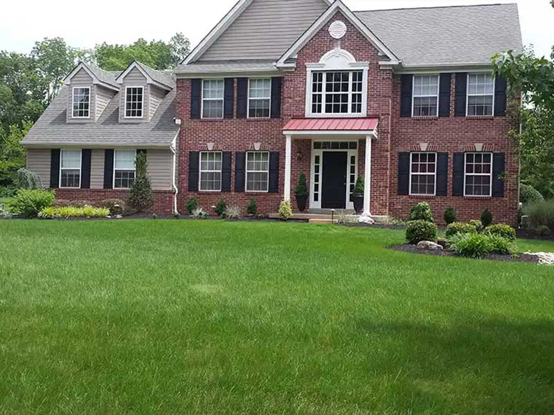 Two-story brick house with black shutters, white trim, and a red metal porch roof, surrounded by a well-maintained green lawn and landscaped bushes.