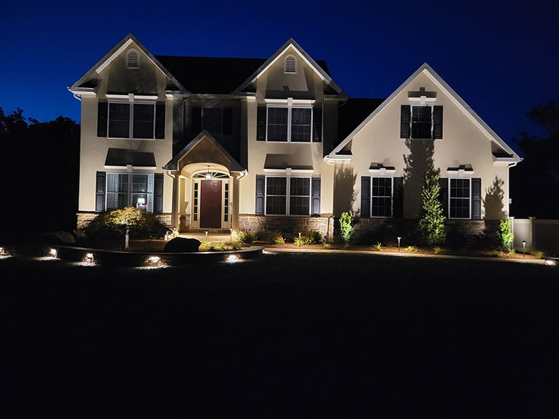 A two-story house with exterior lights illuminating the front entrance, windows, and landscaping at night, set against a dark blue sky.