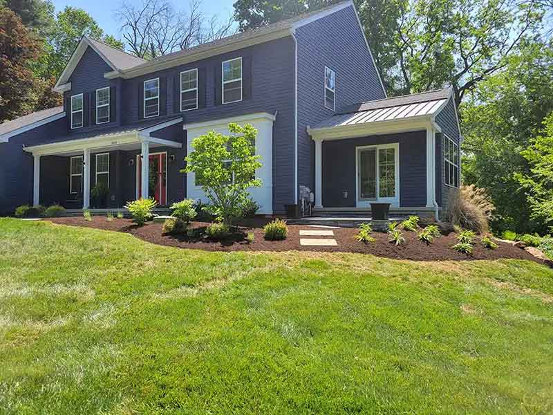 Two-story blue house with white trim and a red door, surrounded by neatly landscaped garden beds and green grass under a sunny sky.