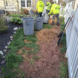 Three workers in yellow jackets clean up a narrow dirt and mulch path beside a white fence, with trash cans and rakes nearby. Green grass and a garden are visible on either side of the path.